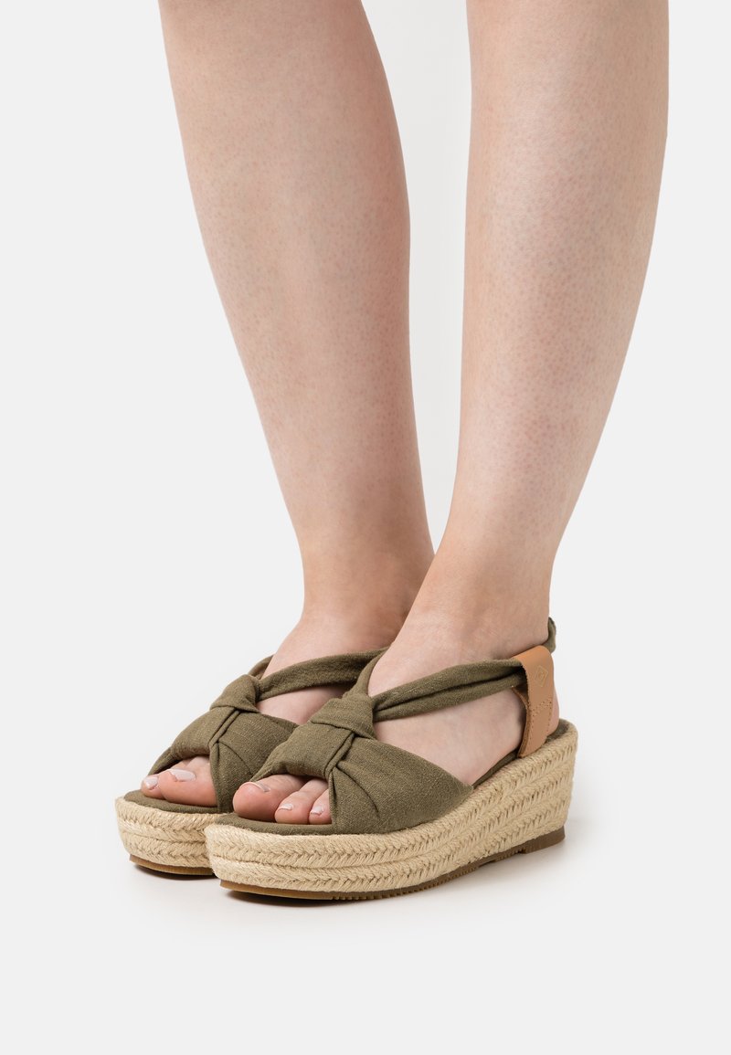 Olive green espadrille sandals with knotted fabric straps, a woven jute platform, and a natural rubber sole. Adjustable ankle strap detail.