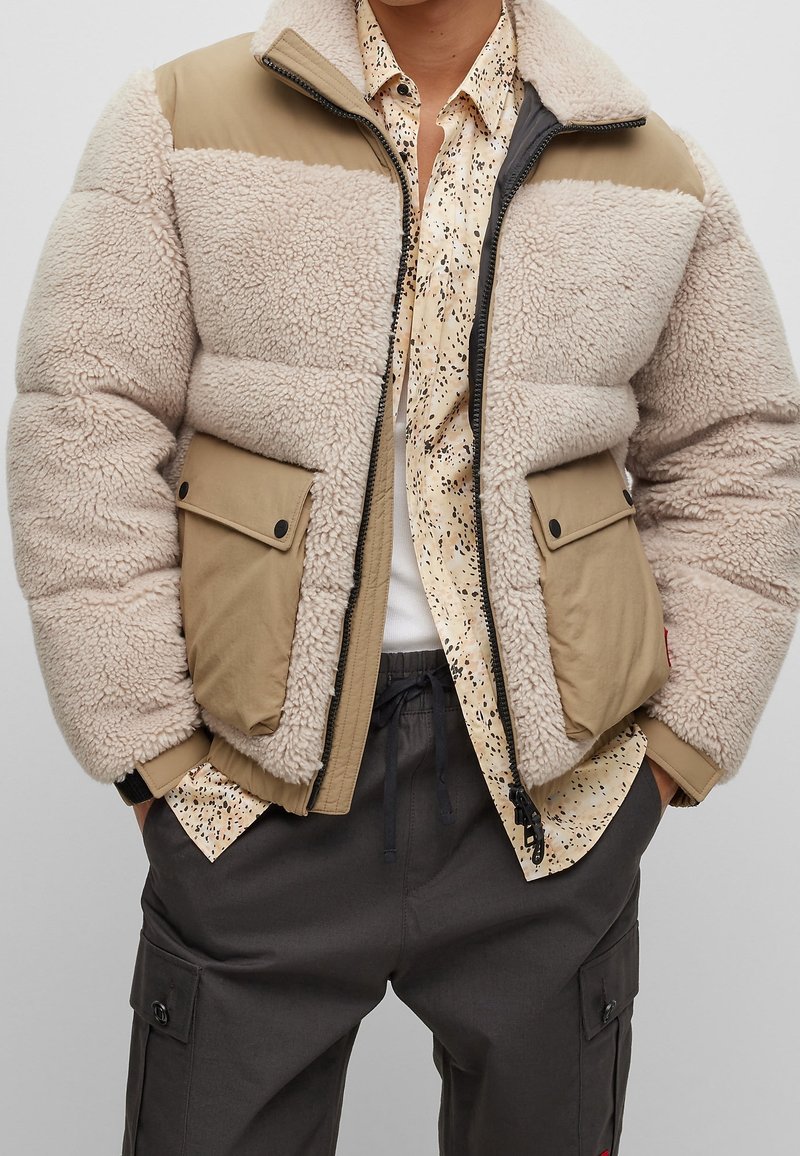 Personne portant une veste sherpa polaire beige et marron clair sur une chemise crème à motifs et un pantalon cargo gris foncé, mains dans les poches.
