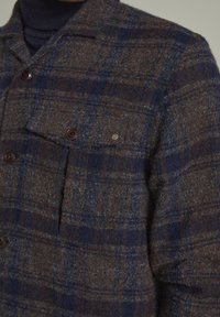 Brun och blå rutiga ullskjortjacka med knappstängning, inklusive en bröstficka och textilstruktur. Har bruna knappar och en logotypaccent.