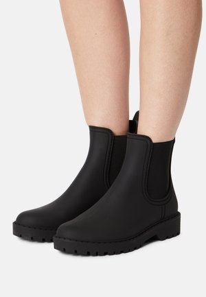 Bottines noires à chevilles avec panneaux latéraux élastiques et semelles épaisses portées sur des jambes nues sur un fond clair uni.