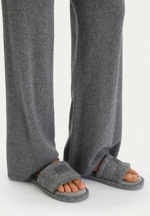 Stopy w szarych miękkich kapciach z wyściełanymi paskami, w połączeniu z luźnymi szarymi spodniami na jednolitym jasnym tle.