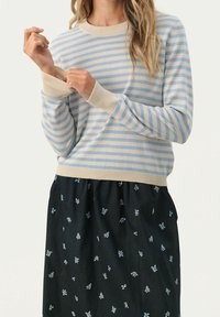 Pull rayé bleu clair et beige avec col rond et poignets côtelés, associé à une jupe noire ornée de petites impressions florales bleues.