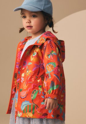 Malé dieťa s modrou čiapkou a jasne oranžovým pršiplášťom s farebnými kvetinovými a hmyzími vzormi stojace na béžovom pozadí.