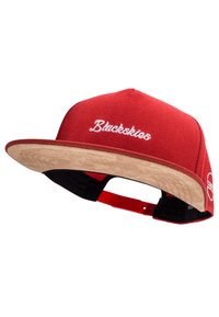 Red cap with a flat tan visor, featuring white embroidery and a red adjustable strap. Textured fabric with contrasting color details.