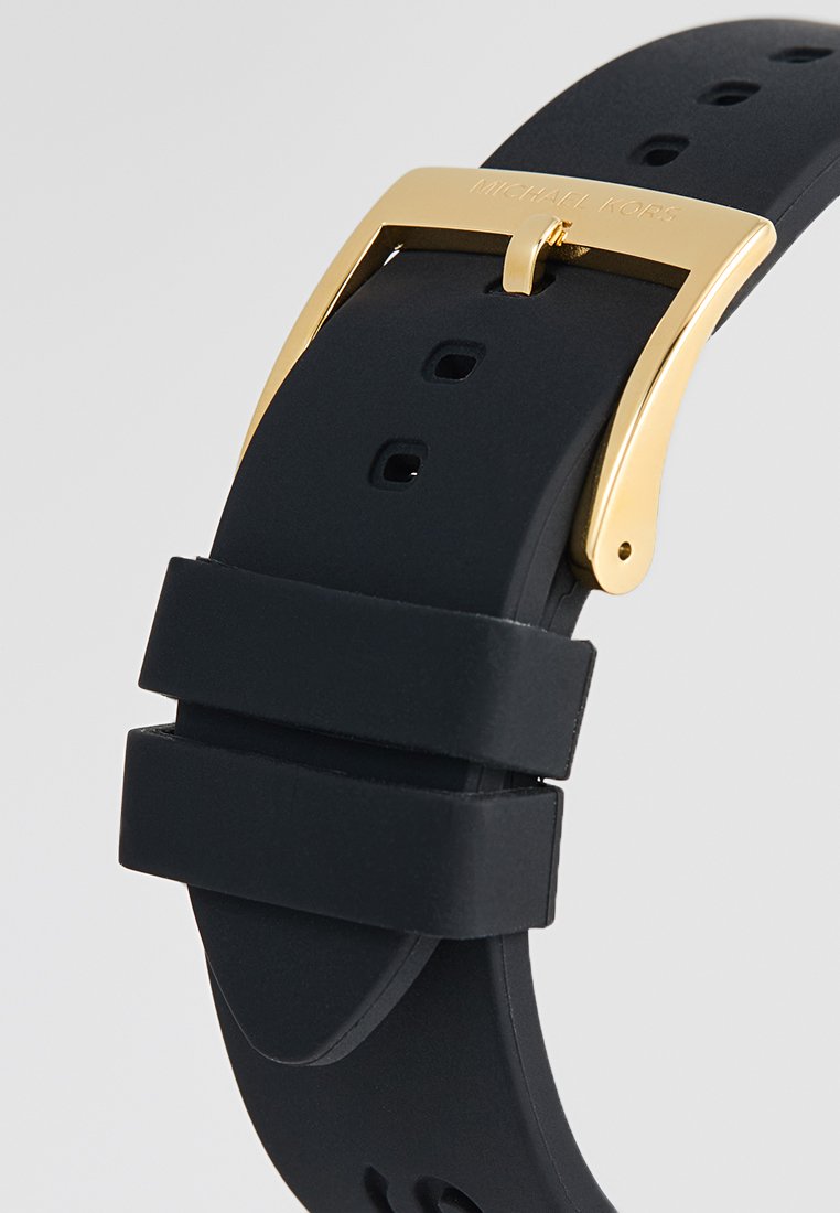 Correa de reloj de silicona negra con hebilla de metal dorado. Presenta múltiples agujeros de ajuste y una textura suave y mate. Detalle de logo grabado.