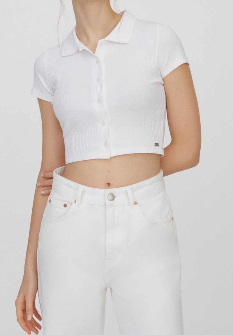 Femme portant une chemise blanche courte à manches courtes, boutonnée avec col, et un jean blanc taille haute, debout avec un bras dans le dos.