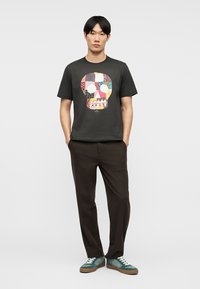 Άνδρας που φοράει μαύρο T-shirt με πολύχρωμο σχέδιο κρανίου σε πατς, σκούρο καφέ παντελόνι και πράσινα αθλητικά παπούτσια, στέκεται με τα χέρια στις τσέπες.