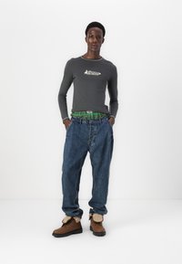 Γκρι μπλουζάκι μακρυμάνικο με λογότυπο, πράσινη καρό ζώνη, φαρδιά μπλε τζιν παντελόνια και καφέ μπότες με μπεζ λεπτομέρειες.