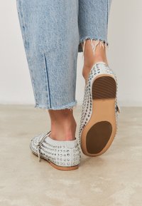 Stopy w jasnoszarych wsuwanych butach z ćwiekami i przyciętych niebieskich dżinsach o postrzępionych nogawkach stoją na gładkiej betonowej podłodze.