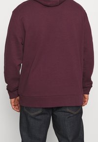 Sudadera marrón con capucha ajustable, puños acanalados y dobladillo inferior, combinada con jeans de mezclilla oscuros que presentan un sutil detalle de costura.