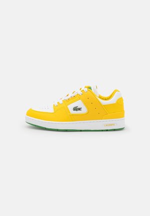 Hvid og gul Lacoste lave sneakers med grønt krokodillelogo, hvide snørebånd og grøn såldetalje på grå baggrund.