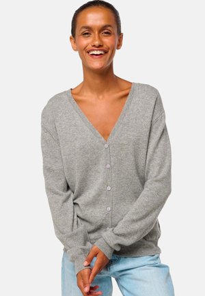 Cardigan en tricot gris avec un col en V, des manches longues et six boutons violet clair. La texture paraît douce et décontractée. Porté par-dessus un jean bleu clair.