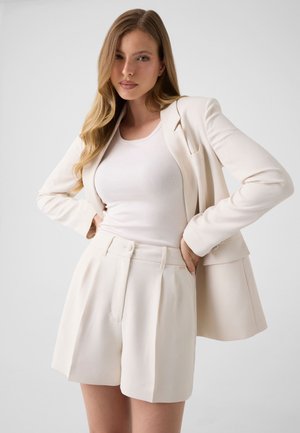 Femme aux longs cheveux blonds portant un blazer crème, un débardeur blanc et un short crème taille haute, posant avec les mains sur les hanches sur un fond gris.