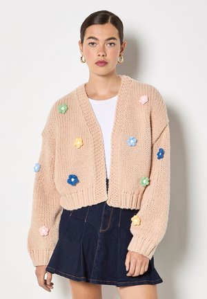Cardigan court tricoté de couleur rose clair avec des appliqués floraux colorés. Il présente un col en V et des manches larges avec des poignets côtelés.