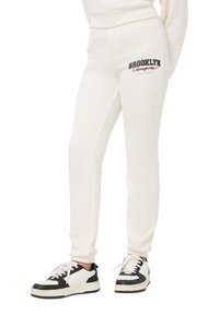 Kremowe dresowe spodnie z dopasowanym stanem, prążkowanymi mankietami u kostek oraz grafiką "BROOKLYN Champions" w czerni i czerwieni na udzie.