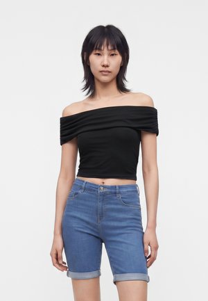 Jeune femme aux cheveux courts et foncés portant un crop top noir épaules dénudées et un short en jean bleu retroussé, debout sur un fond uni.