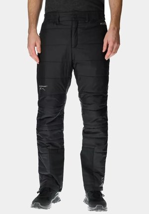 Homme portant un pantalon d'extérieur noir matelassé et des chaussures de randonnée noires, debout devant un fond clair uni.