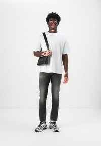 Svart crossover-veske med preget logo, hvit kortermet skjorte, grå slitte jeans og svart-hvite treningssko.