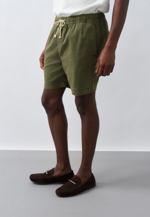 Άνδρας που φοράει χακί σορτς με κορδόνια και σκούρα καφέ μοκασίνια, στέκεται απέναντι σε απαλό γκρι φόντο.