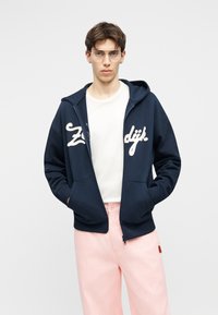 Mladić s prozirnim naočalama, tamnoplavom duksericom na patentni zatvarač s bijelim natpisom, bijelom majicom i svijetloružičastim hlačama, stoji nasuprot bijeloj pozadini.