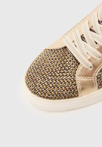 Sneaker with a gold and brown textured upper featuring sparkling studs. It has a beige leather panel and flat cream laces on a white sole.