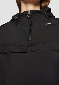 Giacca nera a pullover con zip frontale, cappuccio regolabile con cordino e una texture liscia e opaca. Presenta dettagli in metallo.