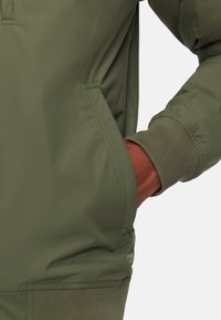 Olijfgroene bomberjack met een gladde, matte afwerking. Voorzien van ribgebreide manchetten en een kangoeroezak, met minimale branding zichtbaar op de zijkant.