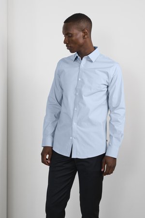 Light blue button-up shirt made from smooth fabric, featuring a collar, long sleeves, and a slim fit design. Paired with dark trousers.