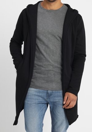 Homme portant un long sweat à capuche ouvert noir, un t-shirt gris et un jean bleu, debout avec une main dans la poche sur un fond uni.