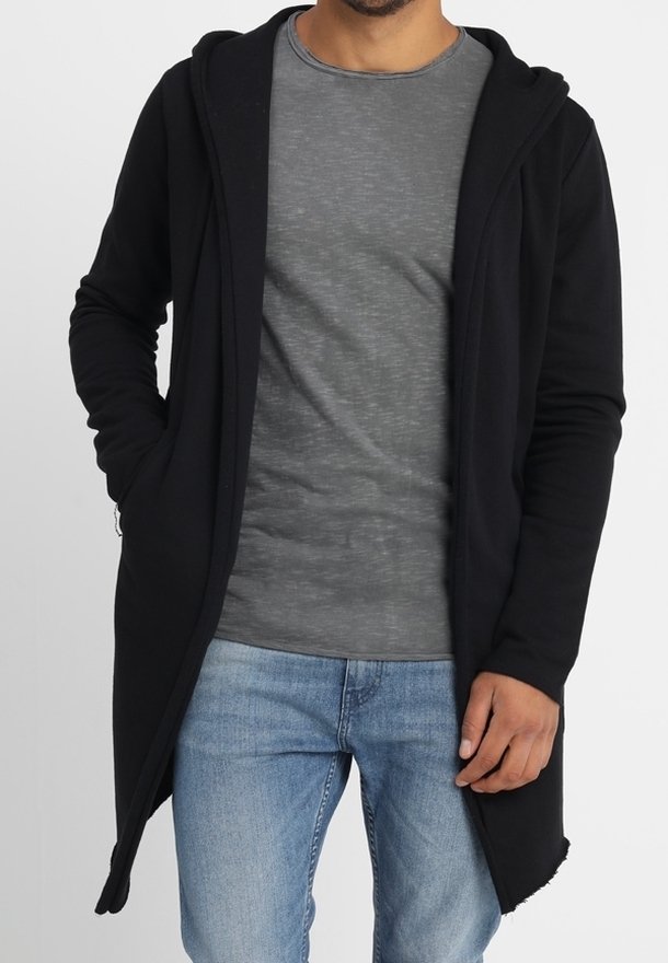 Homme portant un long sweat à capuche ouvert noir, un t-shirt gris et un jean bleu, debout avec une main dans la poche sur un fond uni.
