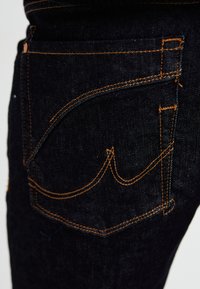 Jeans de ganga escuros com costura laranja. O design notável do bolso traseiro apresenta um padrão de bordado curvo. A textura é suave e resistente.