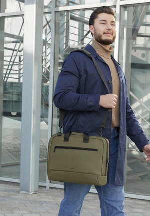 Junger Mann in blauer Jacke und beigem Rollkragenpullover trägt eine olivgrüne Umhängetasche und geht vor einem modernen Glasgebäude.