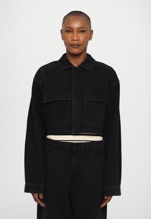 Zwarte cropped denimjack met twee voorzakken, kraag en lange mouwen, gecombineerd met bijpassende denimbroek en een witte binnenlaag.