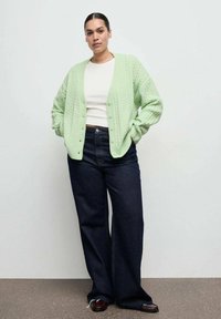 Mäta zelený sveter so zapínaním na gombíky, nosený cez biely top, doplnený tmavomodrými širokými džínsami.