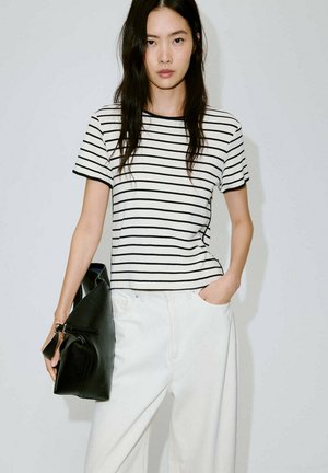 Jeune femme aux longs cheveux foncés portant un t-shirt rayé blanc et noir et un pantalon blanc, tenant un sac à main noir sur un fond uni.