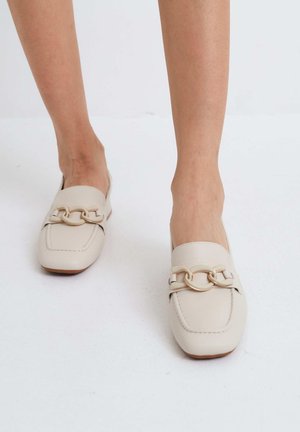 Mocasines de cuero beige con punta cuadrada, con un adorno de cadena dorada en la parte frontal y un acabado liso mate. Diseño de suela plana.