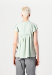 Blusa verde chiaro a maniche corte con schiena arricciata e dettagli ricamati. Tessuto morbido con vestibilità rilassata, abbinata a jeans grigi.