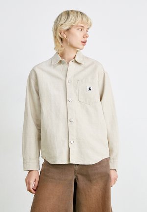 Beige long-sleeve shirt with a button-down collar, featuring a left chest pocket and a logo patch. Smooth texture and loose fit design.