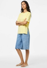 T-shirt di cotone giallo con maniche corte, fessure laterali, abbinata a pantaloni di jeans. I pantaloni sono blu, di lunghezza al ginocchio, con orlo sfrangiato.