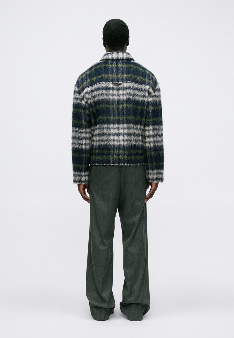 Persona de espaldas vistiendo una chaqueta de cuadro azul, blanca y verde de felpa y pantalones oscuros de corte holgado contra un fondo sencillo.