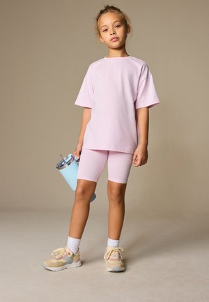 Junges Mädchen steht in einem rosa übergroßen T-Shirt und passenden Radhosen, hält einen hellblauen Becher mit Strohhalm und trägt weiße Socken und Turnschuhe.