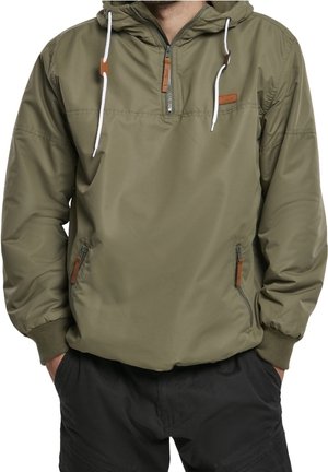 Muž v olivově zelené bundě s kapucí, předním zipem, bílými šňůrkami a zipovanými bočními kapsami, v kombinaci s černými kalhotami.