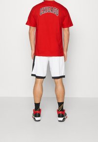 Hombre de pie con la espalda hacia la cámara, vistiendo una camiseta roja con la palabra "CHICAGO", pantalones cortos deportivos blancos y negros, calcetines negros y zapatos deportivos rojos y negros.