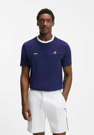 Mężczyzna ubrany w granatowy T-shirt z napisem "BOSS" i tarczą z flagą USA, w połączeniu z białymi szortami ze sznurkiem i napisem "BOSS" po boku.