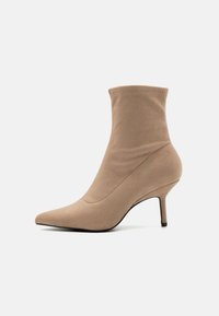 Bottine beige à mi-mollet en suede doux. Elle présente un bout pointu, un talon fin et un design sans couture avec des détails de couture subtils.