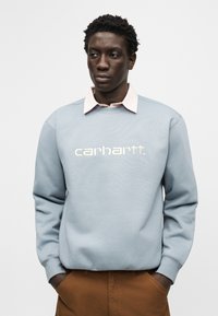 Νέος άνδρας με ανοιχτό μπλε φούτερ Carhartt πάνω από ανοιχτό ροζ πουκάμισο με γιακά και καφέ παντελόνι, με τα χέρια στις τσέπες, κοιτάζοντας προς τα αριστερά.