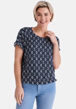 Frau mit kurzem blondem Haar, die ein kurzärmeliges Oberteil mit marineblauem und weißem geometrischem Muster und hellblaue Jeans trägt, lächelt in die Kamera.