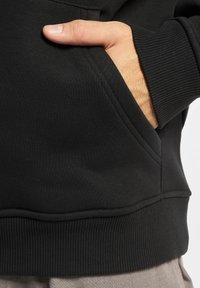 Svart hettegenser med frontlomme, ribbede mansjetter og nederkant, myk tekstur og tettsittende design. Hånd hviler i lommen.