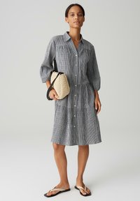 Gestreepte button-up jurk in grijs en wit, met opgerolde mouwen, gelaagd ontwerp en lichte stof. Gecombineerd met een geweven crèmekleurige handtas.
