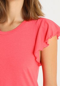 Korálově růžové tričko s krátkými volánkovými rukávy, kulatým výstřihem, vyrobené z jemné, texturované látky. Jednoduchý design bez dalších vzorů nebo akcentů.
